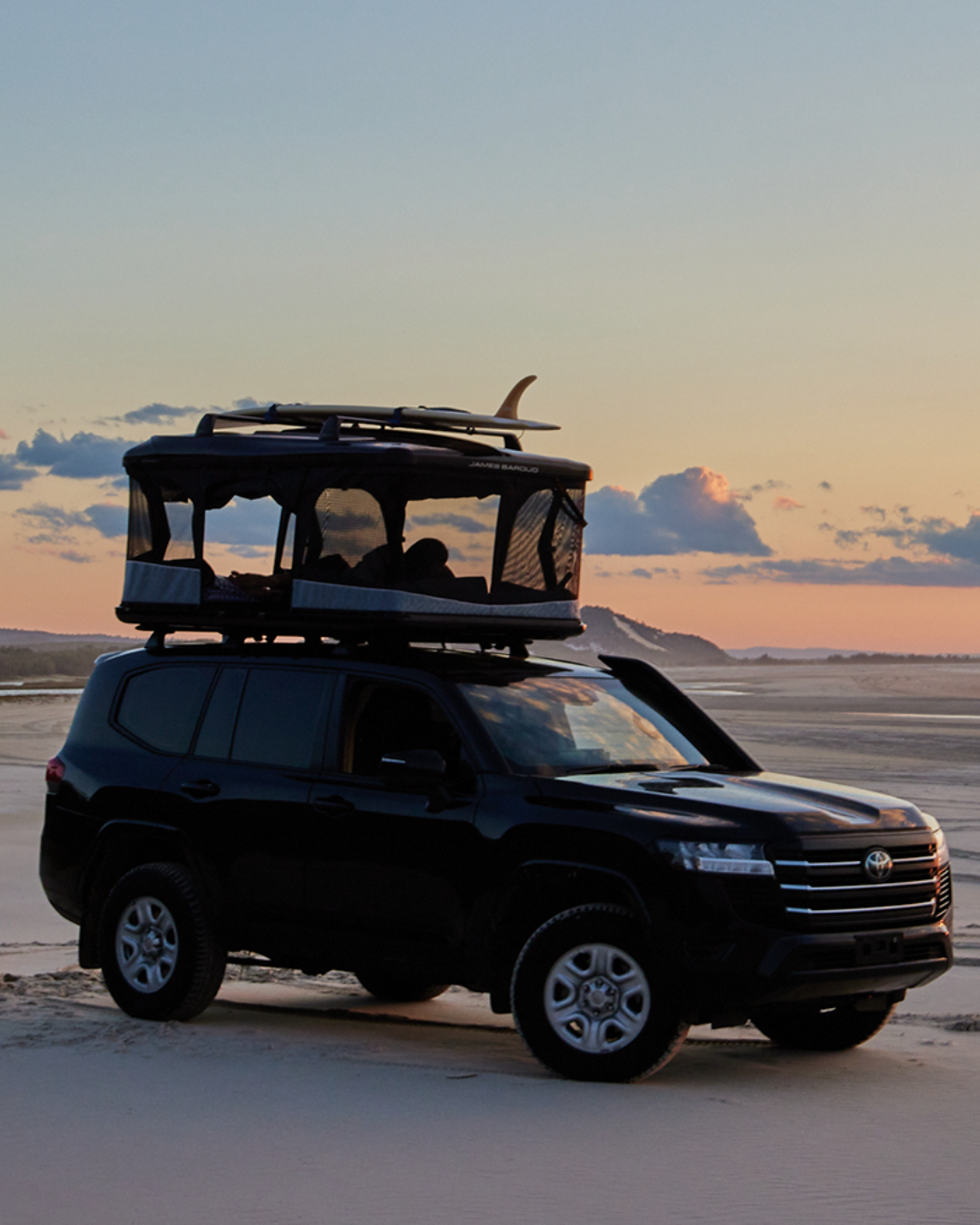 James Baroud Hartschalen-Dachzelt Odyssey schwarz montiert auf einem Offroad-Fahrzeug. Dachzelt ist offen. Person liegt im Dachzelt. und ein Surfbrett wurde auf den Querträgern auf dem Zeltdach montiert.