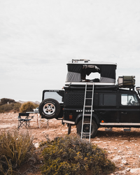 James Baroud Hartschalen-Dachzelt Grand Raid schwarz montiert auf einem Offroad-Fahrzeug. Dachzelt ist aufgebaut. Fenster offen. Mit Leiter Zustieg. Gepäck auf dem Dach.