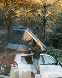 James Baroud Hartschalen-Dachzelt Discovery schwarz montiert auf einem Offroad-Fahrzeug. Dachzelt ist aufgebaut. Mann öffnet Einstieg über die Leiter.