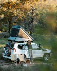 James Baroud Hartschalen-Dachzelt  Discovery montiert auf einem Offroad-Fahrzeug beim Campen. Ein Mann klettert über die Leiter hoch.