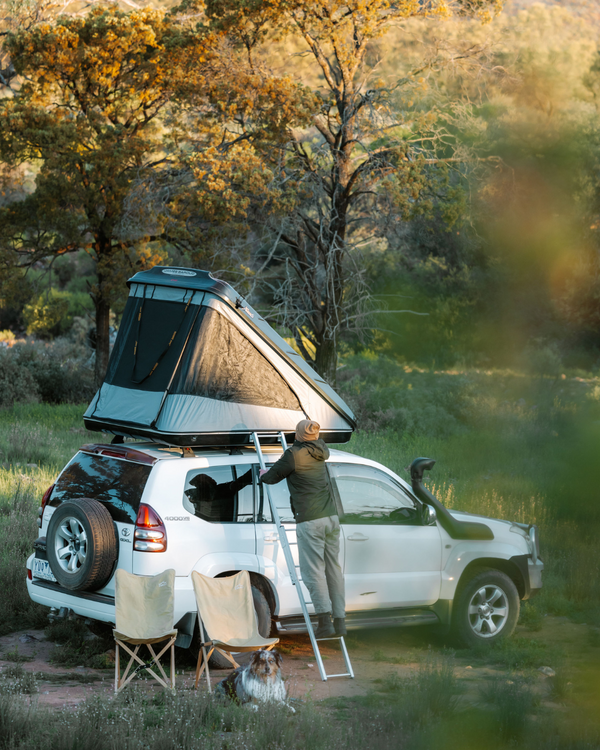 James Baroud Hartschalen-Dachzelt  Discovery montiert auf einem Offroad-Fahrzeug beim Campen. Ein Mann klettert über die Leiter hoch.