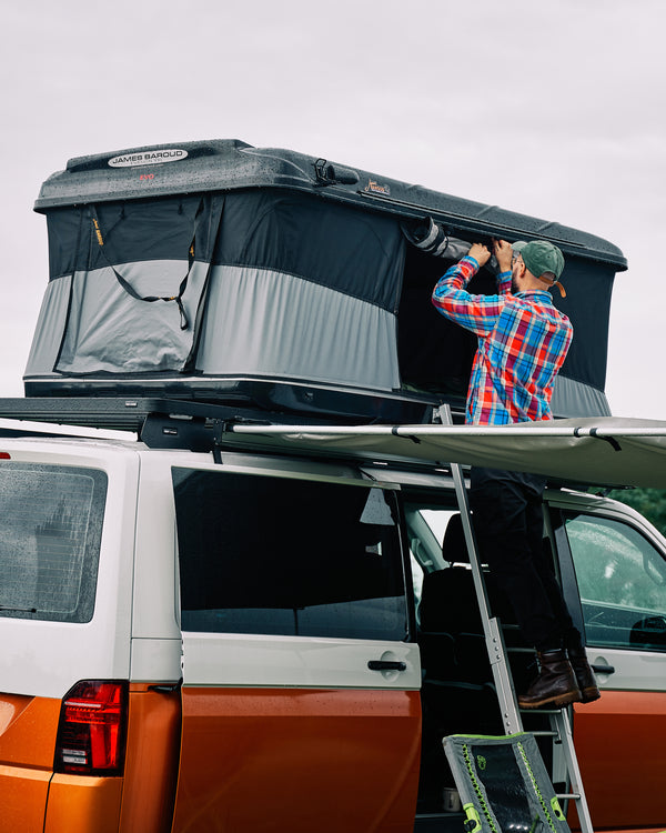 James Baroud GFK Hartschalen-Dachzelt schwarz montiert auf einem Offroad-Fahrzeug. Dachzelt ist aufgebaut. Mit Leiter Zustieg. Mann rollt Eingangsfenster nach oben. Mit Markise.