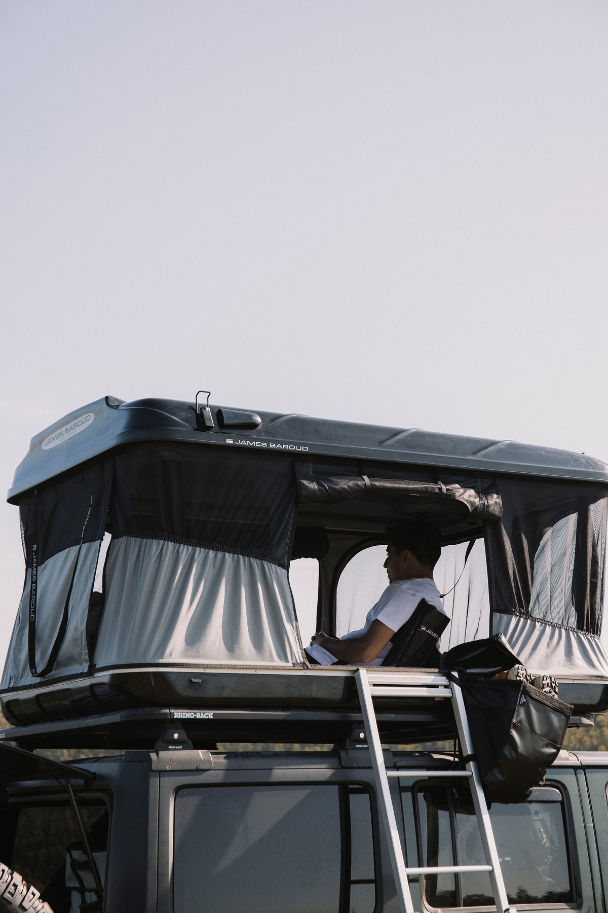 James Baroud Hartschalen-Dachzelt schwarz offen montiert auf einem Offroad-Fahrzeug. Beim Campen. Mit montierter Schuhtasche am Dachzelt. Im inneren sitzt ein Mann im James Baroud klappbaren Stuhl.