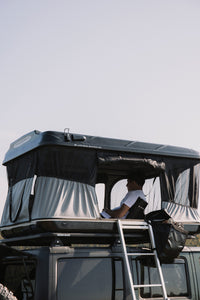 James Baroud Hartschalen-Dachzelt schwarz offen montiert auf einem Offroad-Fahrzeug. Beim Campen. Mit montierter Schuhtasche am Dachzelt. Im inneren sitzt ein Mann im James Baroud klappbaren Stuhl.