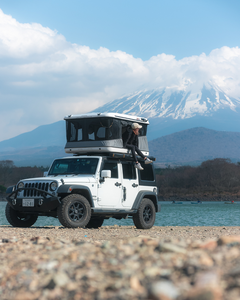 James Baroud Hartschalen-Dachzelt Evasion weiß montiert auf einem Offroad-Fahrzeug. Dachzelt ist aufgebaut. Frau sitzt am Einstieg.